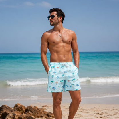 A shirtless man wearing light blue swim trunks with a palm tree pattern stands on a sandy beach with the ocean in the background.