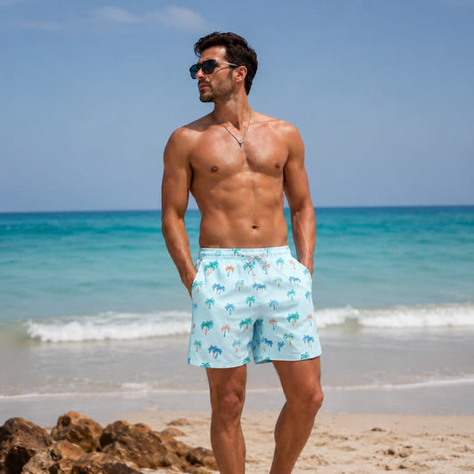 A shirtless man wearing light blue swim trunks with a palm tree pattern stands on a sandy beach with the ocean in the background.