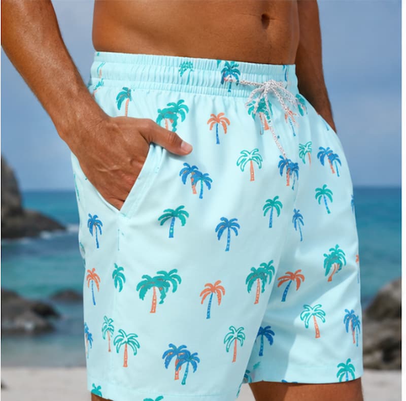 Men's swim trunks with a light blue background and a green and orange palm tree pattern, shown on a model at a beach.