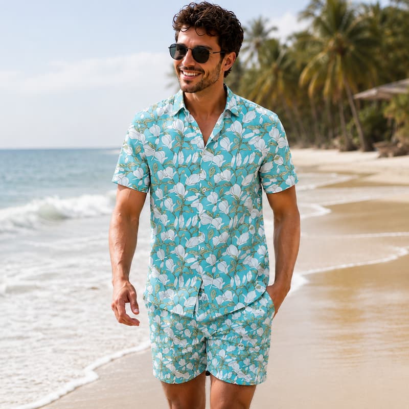 A man wearing a matching turquoise and white tropical floral print short-sleeve button-up shirt and shorts set, walking on a sandy beach with palm trees and the ocean in the background.