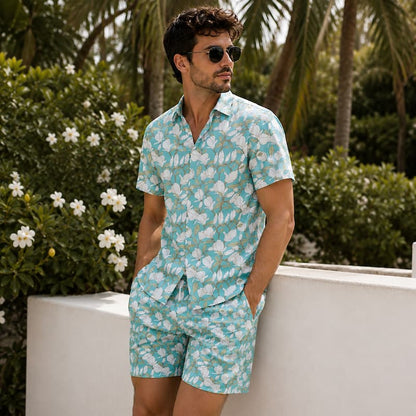 A man wearing a light blue and white tropical floral print short-sleeve button-up shirt and matching shorts, standing outdoors against a white wall with green foliage and palm trees in the background.