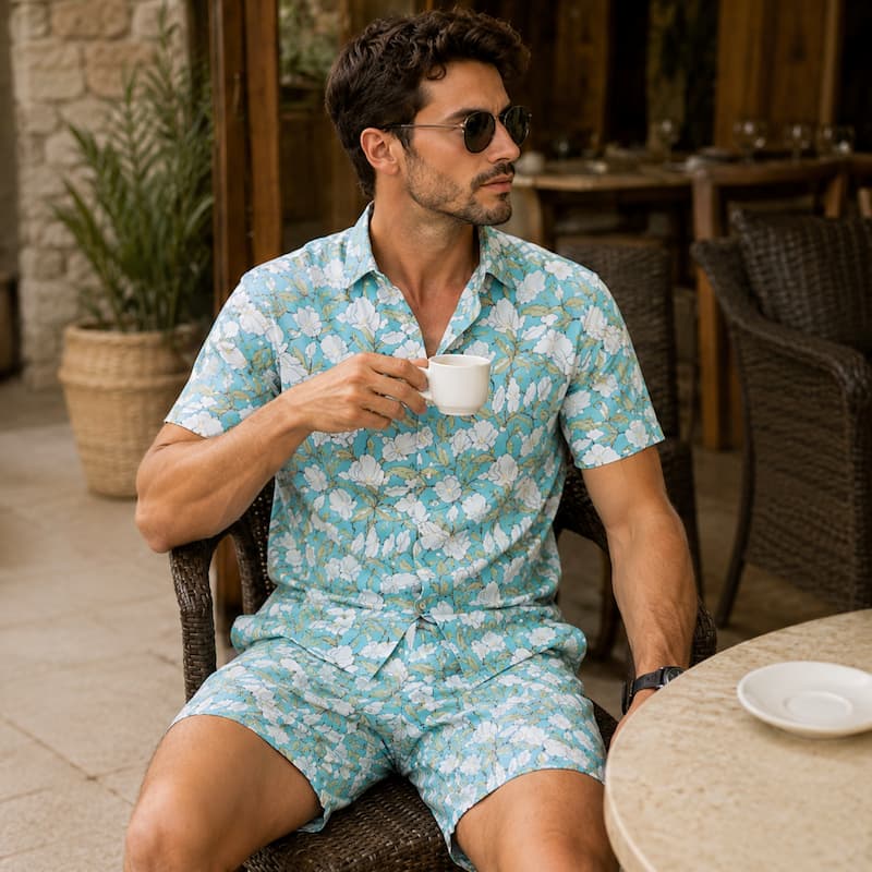 A man wearing a light blue and white floral print short-sleeve button-up shirt and matching shorts, sitting at an outdoor cafe table and holding a white coffee cup.