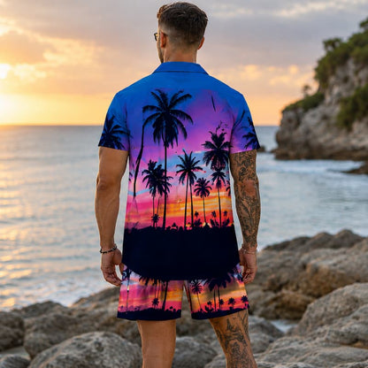 A man stands on a rocky beach at sunset wearing a short-sleeve button-up shirt and matching shorts with a colorful tropical palm tree sunset print. The shirt and shorts feature a gradient design transitioning from blue and purple at the top to pink, orange, and black silhouettes of palm trees near the bottom.